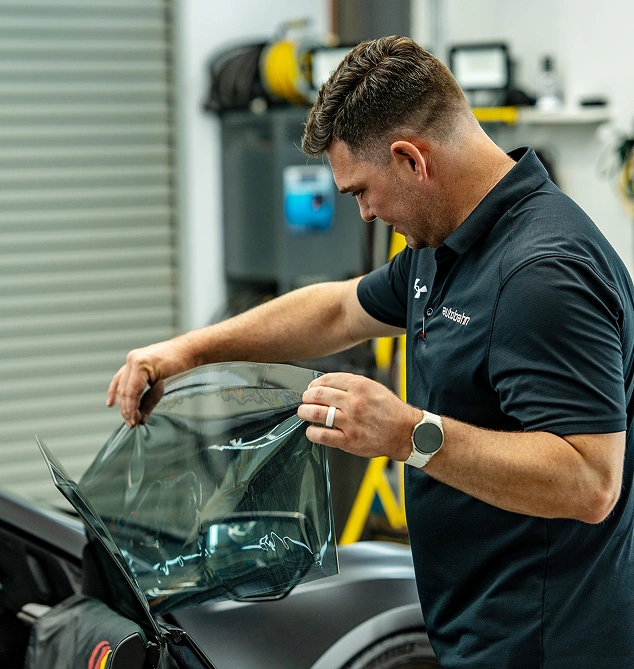 Man in an autobahn polo shirt applying a sheet of window tint film to a car window inside a workshop.