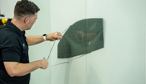 Man in a black polo shirt applying window tint film to a detached car window mounted on a vertical surface.