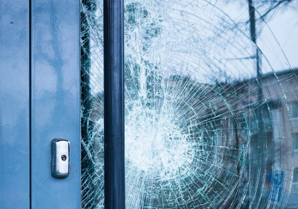 Close-up of a large circular crack in a glass door, with spiderweb-like fractures indicating impact damage, next to a metal handle and frame.