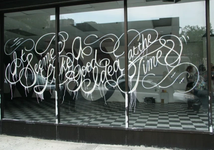 Large graffiti-style white script reading “It seemed like a good idea at the time” painted across the windows of an empty storefront with checkered floors.