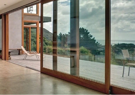 Large sliding glass doors in a modern home reflect an outdoor deck with lounge chairs, trees, and an ocean view in the distance.